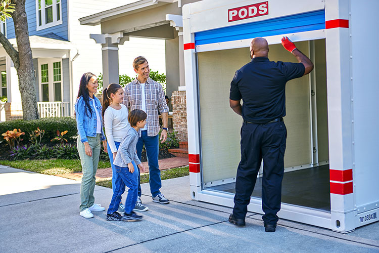 A family (mom, dad, and two kids) looks on as their PODS delivery driver closes the door to the moving and storage container in their driveway. 
