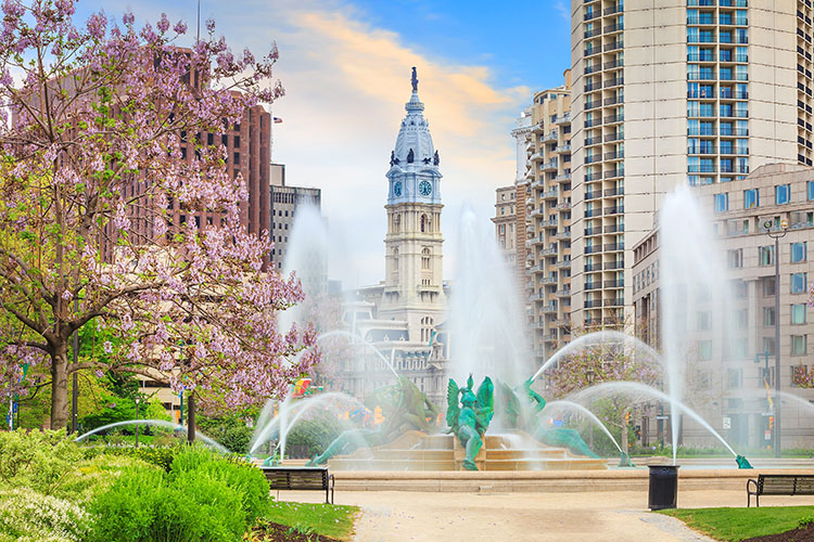 Philadelphia’s Swann Memorial Fountain in the springtime. Blooming trees and greenery are in the foreground, with tall buildings surrounding Logan Square in the background. 