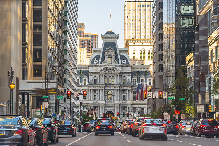 Heavy traffic, most likely during rush hour, in front of Philadelphia’s City Hall. There are four lanes of bumper-to-bumper with tailights illuminated. 