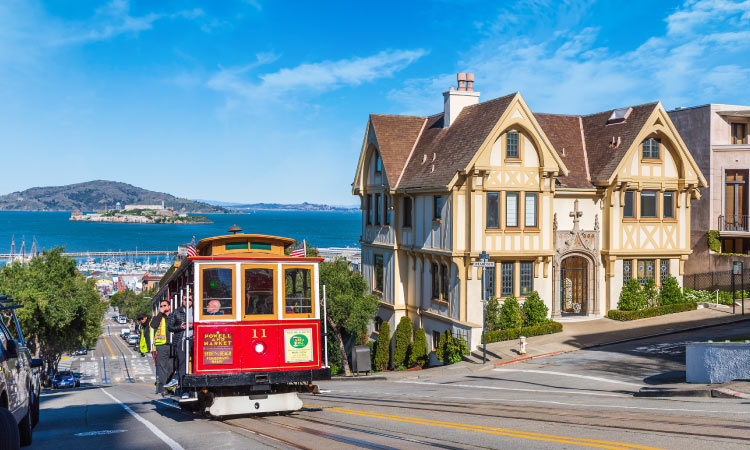  A cable car in San Francisco climbs a steep road past historic residential buildings, with a view of the Bay in the distance.