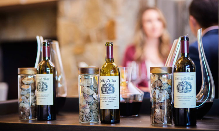 Three bottles of Napa Valley wine are displayed on a table beside decorative glass jars in a restaurant. A couple is dining out of focus in the background.