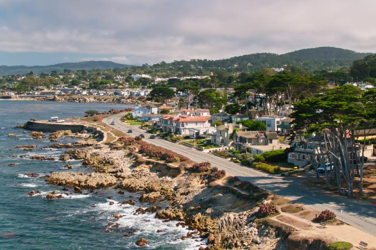 Aerial view of a coastal road in Pacific Grove, California. The residential side of the road is lined with high-end homes and shade trees, and the coastal side is rocky with a steep drop-off.