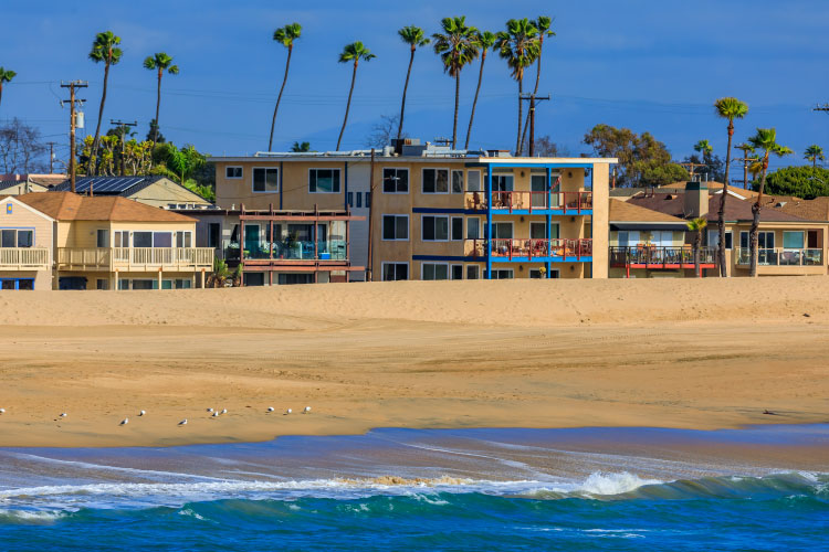 View of beach houses and condos in Seal Beach, California, seen from the water. The Pacific is a bright blue, and the sand on the beach looks soft and fine. Tall, skinny palm trees poke up from behind the beachfront buildings.