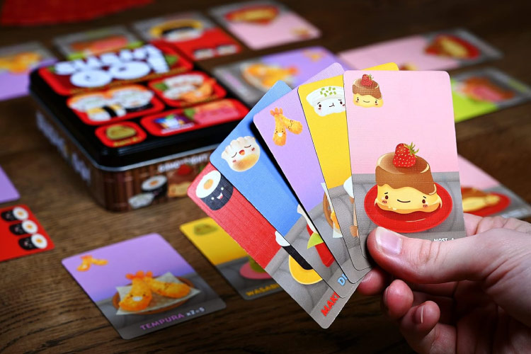 Close-up view of a hand holding five cards from the Sushi Go! card game. More cards and the game tin are seen in the background on a wooden table. 