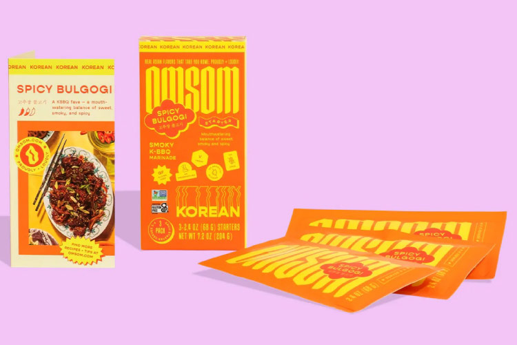 Three Omsom Spicy Bulgogi sauce packets and the box they come in are set against a light pink background.