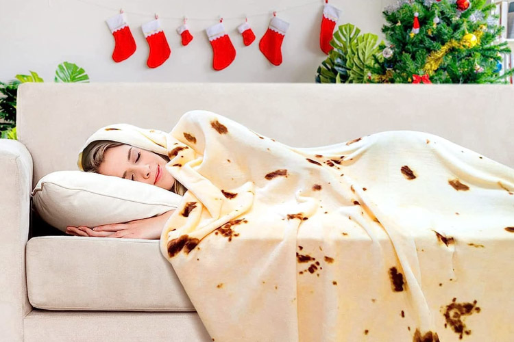 A woman is napping happily on her couch, covered in a blanket that looks like a tortilla. Behind the couch is a Christmas tree and stockings.