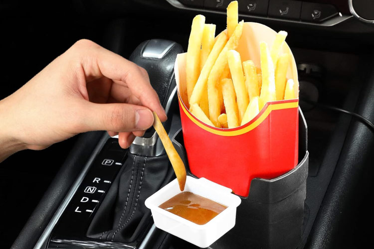 Close-up of a hand dipping a french fry in a sauce container. The french fry box and sauce container are held in a specialized cup holder attachment in a car’s center console. 
