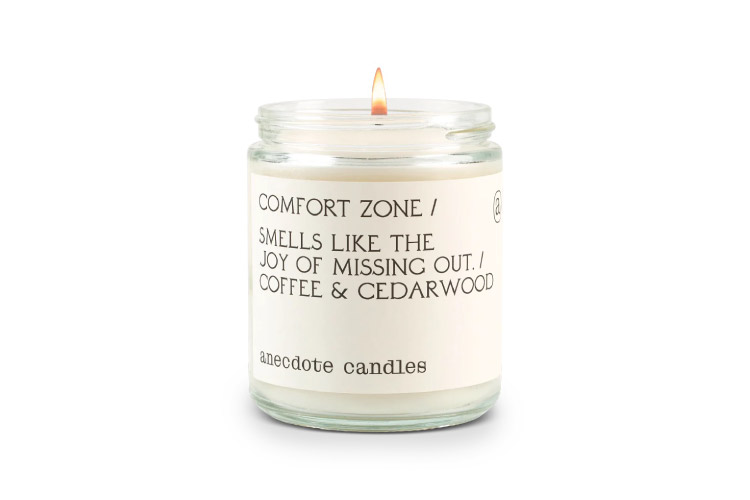 A single Anecdote Comfort Zone candle. The lit candle is in a clear glass jar and the label on the jar reads, “Comfort Zone / Smells like the joy of missing out. / Coffee & Cedarwood.” 