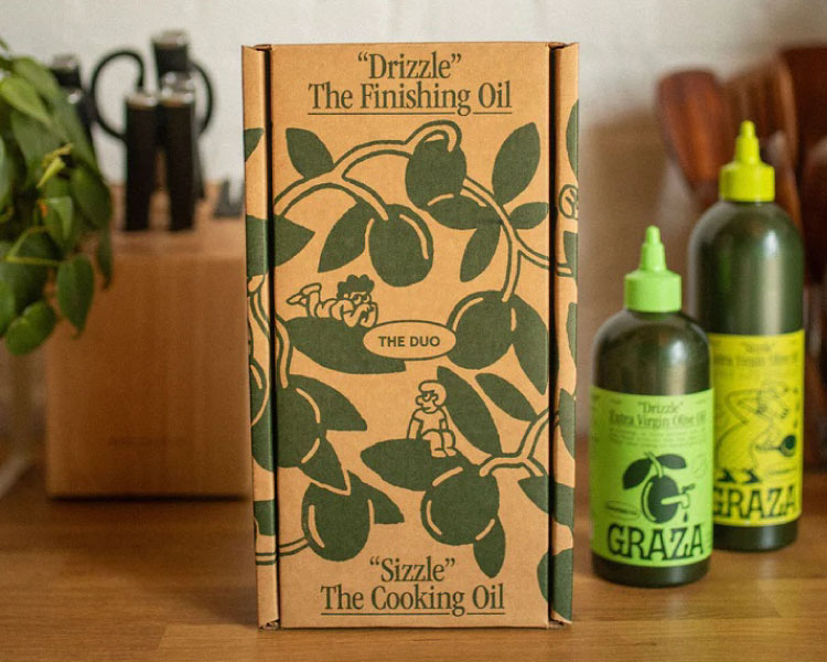 A kitchen countertop with the Graza Olive Oil Gift Set. The two bottles of olive oil are arranged beside the cardboard box they came packed in.