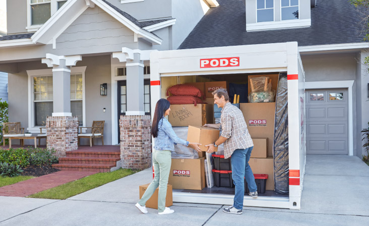 A mature couple is working together to load moving boxes into a PODS portable moving container in their driveway. The container is nearly full and is neatly packed with boxes, plastic bins, pillows, a stool, and a mattress.