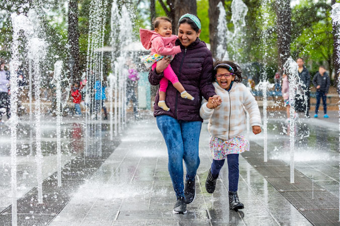 A mother is carrying her toddler and holding the hand of her young daughter as they happily run through a splash pad in Beaverton, Oregon — one of the best Portland suburbs.