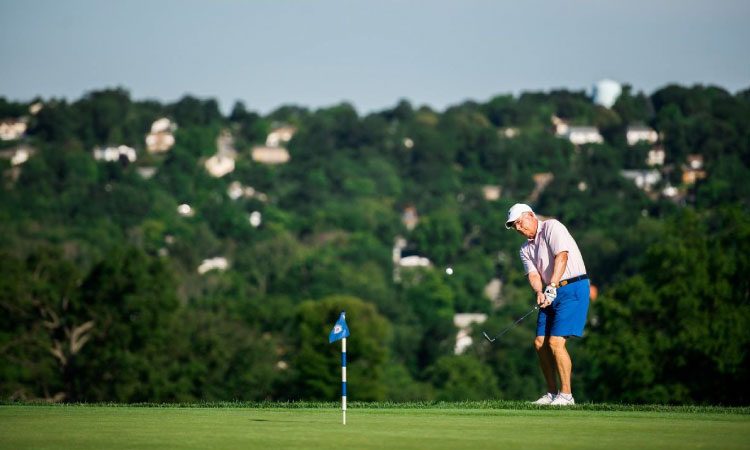 A senior man putts at the Montclair Golf Club in Montclair, New Jersey. Behind him are lush hills filled with residential homes in the area surrounding the golf club.