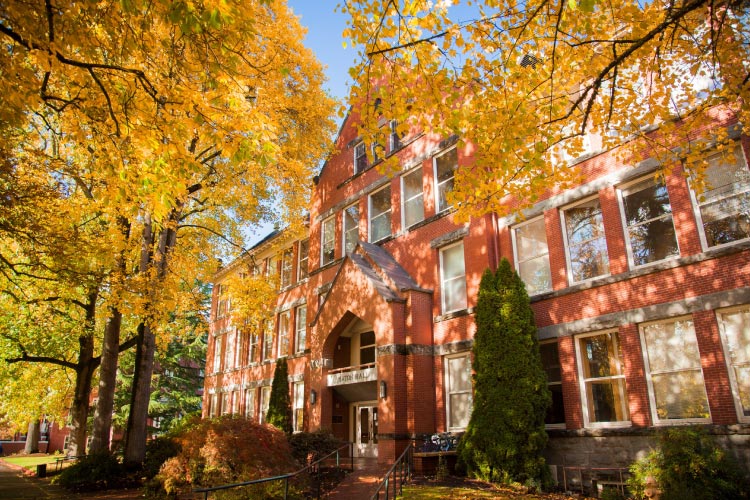 The red brick Eaton Hall on the campus of Willamette University in Salem, Oregon. The building is nestled among tall trees that have begun to change color in the fall.