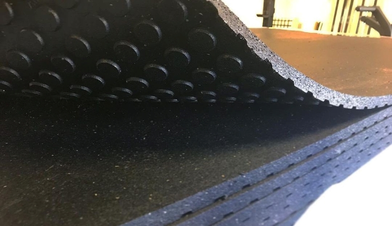 A stack of rubber garage gym floor mats with textured bottoms to keep them from slipping. The top mat is being lifted up to reveal the texture beneath. Behind the mats, parts of various exercise machines are visible.