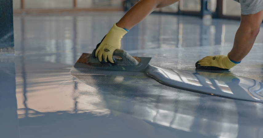 Close-up view of a person carefully smoothing out the first layer of a blue epoxy finish on a garage floor. The person is wearing protective gloves and is using a notch trowel to move the epoxy around and get a flat finish.