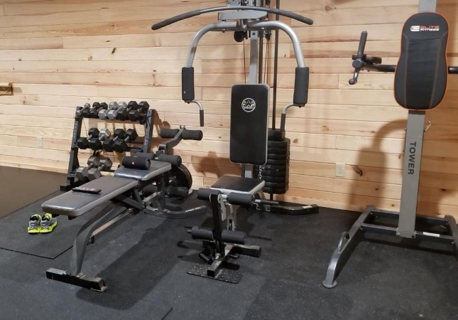 A garage gym with rubber gym flooring beneath free weights and a variety of exercise machines. The walls of the garage are covered with horizontal wooden planks giving it a finished look.