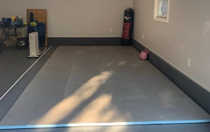Gray and blue interlocking foam floor mats on a garage gym floor. There is a single punching glove resting on the edge of the mats and a punching bag leaning up against the corner of the garage. On the other side of the mats, various household items are arranged in no particular order.