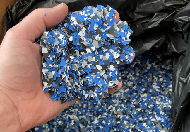 A close-up of a hand holding a bunch of blue, gray, and black decorative vinyl flakes that will be used to break up the color of an epoxy floor. In the background is a large black plastic bag filled with more of the same flakes. 