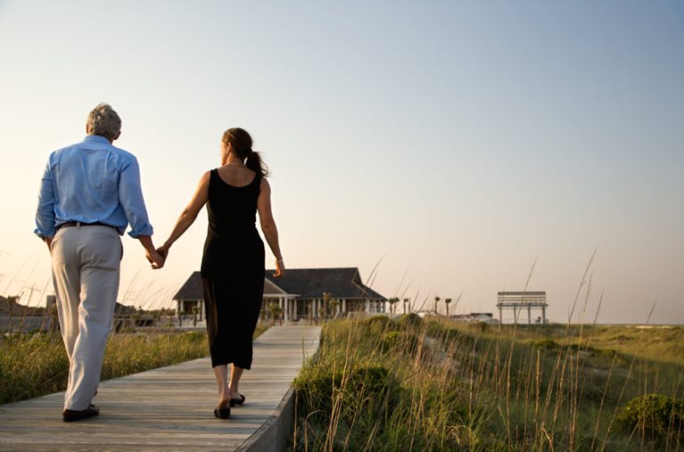 A recently retired couple is walking hand-in-hand up a boardwalk on a beach in North Carolina. 