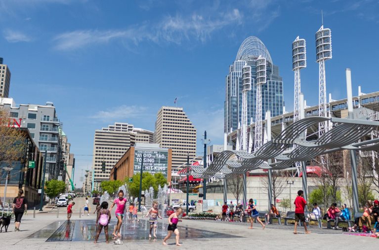 Dozens of locals enjoy a sunny day at a city park in Cincinnati, Ohio, with a view of the city skyline in the background.