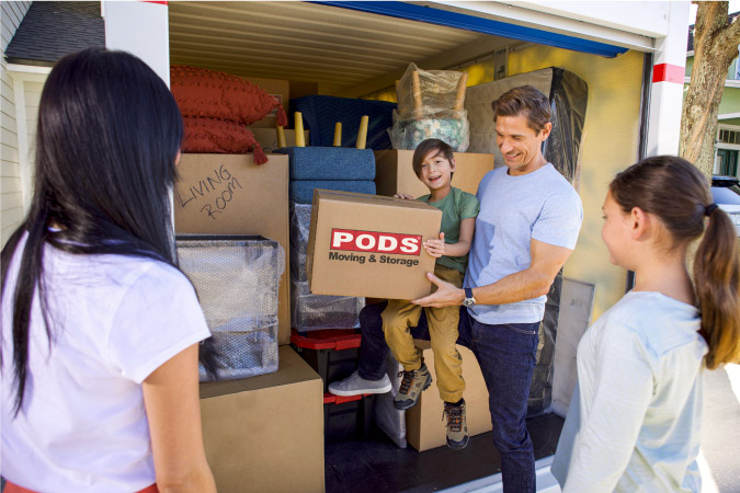 A family of four are standing at the door of their PODS portable storage container, which is fully loaded with furniture and moving boxes. 