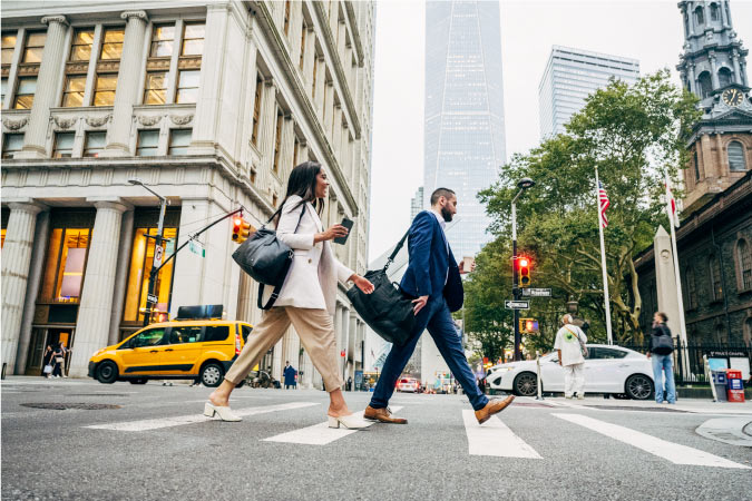 A man and woman in business attire cross Fulton on Broadway in NYC’s Financial District. 