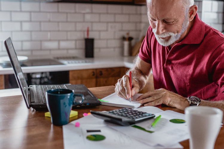 A senior man is sitting at his kitchen table with his laptop, notepad, and calculator, trying to decide is North Carolina a good place to retire for him?