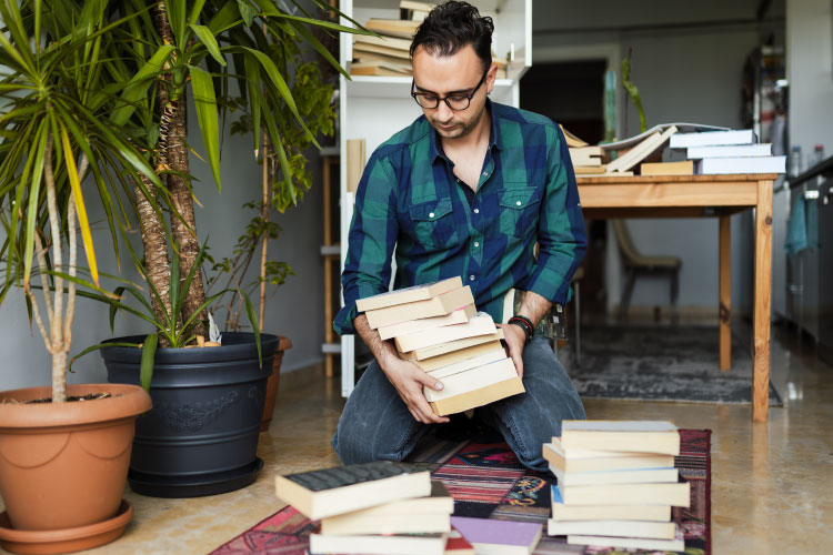A man is kneeling on the floor of his living room, going through stacks of books. He’s decluttering before having hardwood floors installed.