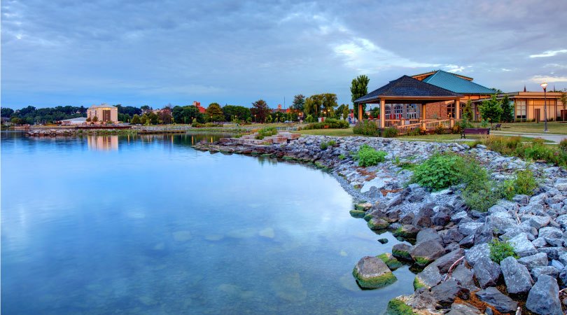 The sun has just set along the shore of Seneca Lake in Geneva, New York. The waters of the lake are still and clear. They’re reflecting the sky as well as the buildings close to shore. In the distance, the tops of the downtown buildings peek out above the trees