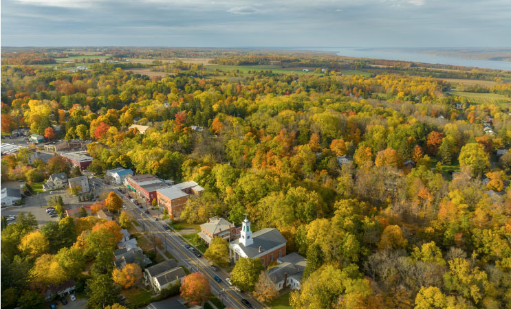 Aerial view of the small town of Trumansburg, New York. It’s late summer and some of the trees have already started to change their leaves. The landscape is heavily wooded but relatively flat, making it possible to see Cayuga Lake in the distance.