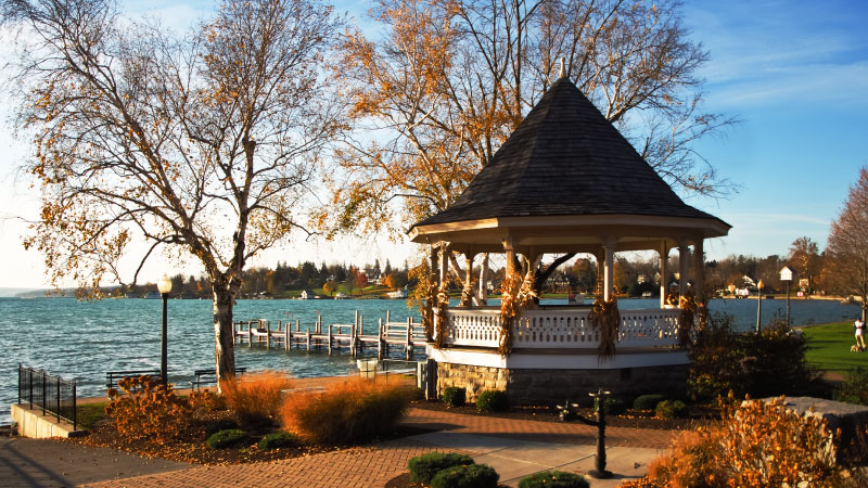 A beautiful autumn day on the shores of Skaneateles Lake in Skaneateles, New York. There’s a lovely gazebo and a wooden dock in the foreground and, on the distant shore, there are residential homes