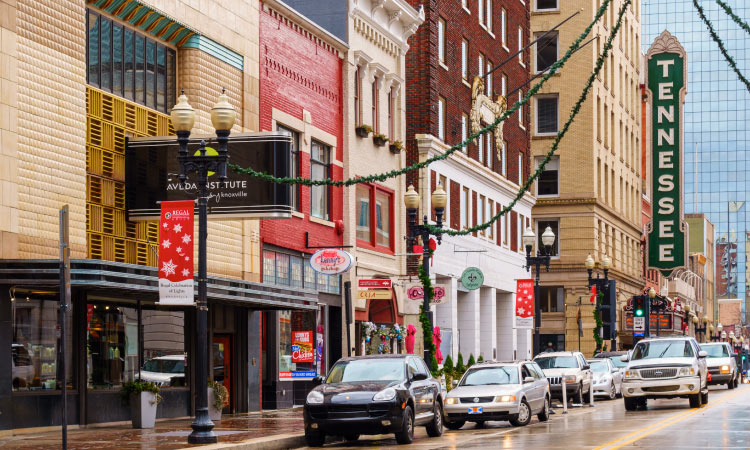 Street-level view of Downtown Knoxville on a rainy day. Cars are traveling down the streets past charming storefronts and the light poles are decorated for the winter holiday season. A large sign with the word “TENNESSEE” is featured on the side of a large building in the background. 