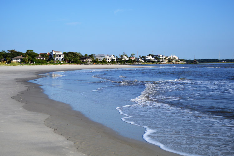 View down the beach of beachfront homes in St. Simons Island, Georgia. The sand is a light cream color, and the water is lapping gently against the shore.