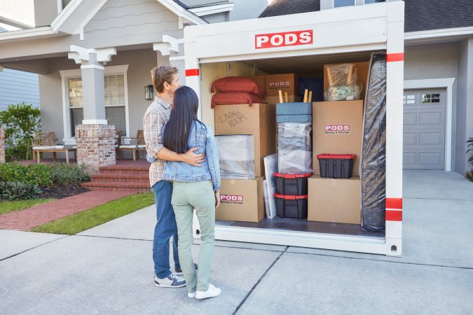 A couple is standing arm in arm in front of the PODS portable moving container in their driveway. The container is neatly packed with a mattress, chairs, moving boxes, and miscellaneous household items.