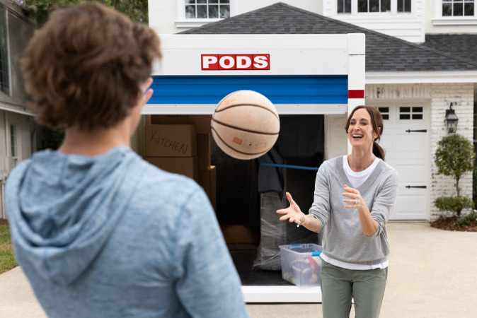 A mother and her teenage son are tossing a basketball back and forth while taking a break from their move to Cincinnati. There’s a partially loaded PODS portable moving and storage container in the driveway behind them.