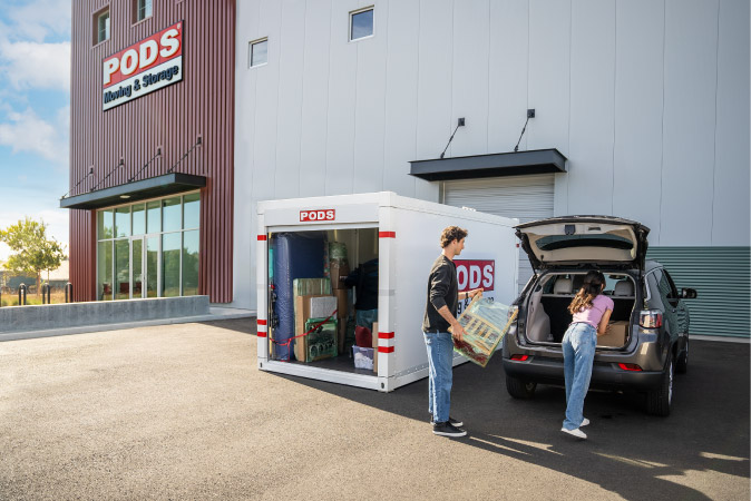 A couple is pulling a few things out of their PODS portable storage container and placing them into the trunk of their car outside a PODS storage center.