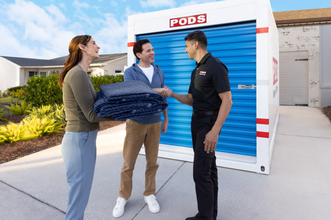 A couple is talking with a PODS driver after he dropped off their portable PODS moving container. The woman is holding moving blankets and the man is shaking hands with the driver.