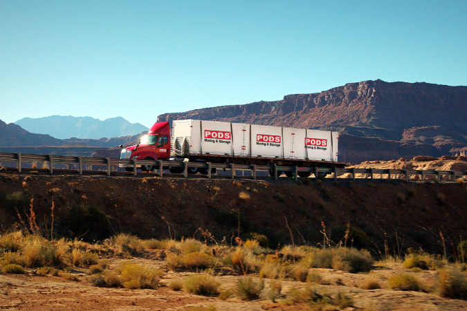 A PODS truck is transporting three PODS portable moving containers along an interstate past breathtaking western vistas.