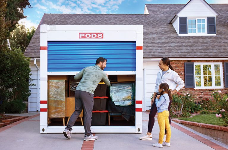 A man is closing the blue, roll-down door on a loaded PODS portable moving container as his wife and young daughter watch from nearby.