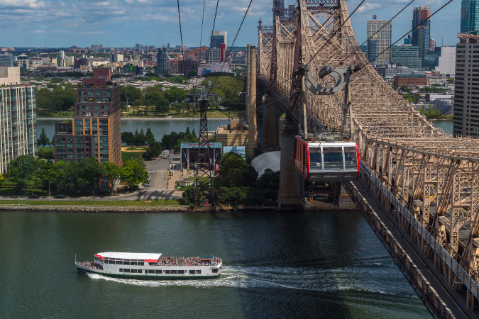 Aerial view of the Roosevelt Island Tram beside Queensboro Bridge in NYC, with Roosevelt Island in the background and a ferry in the river below.