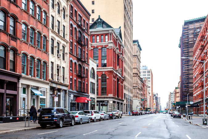 A wide one-way street in NYC’s Tribeca neighborhood features red brick buildings with residentials levels and commercial storefronts.  