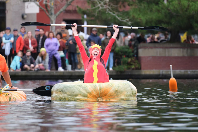 A woman dressed as a hotdog is sitting in her pumpkin boat, holding her paddle high in the air in celebration during the annual Pumpkin Regatta Festival.