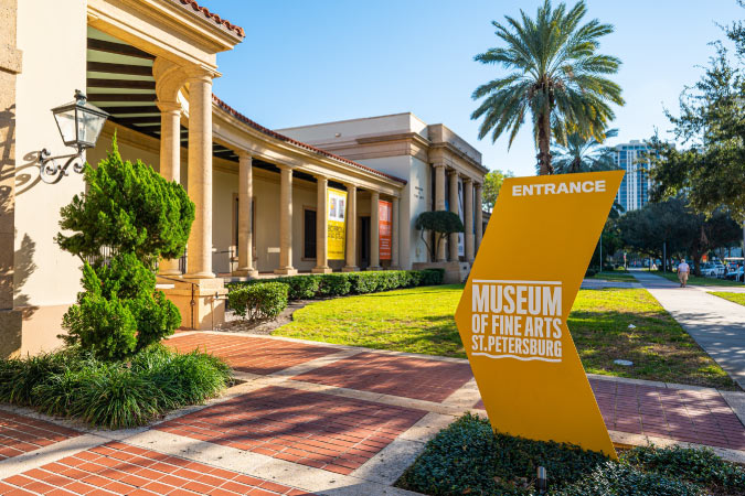 On a sunny day in St. Petersburg, Florida, Roman-inspired columns, pruned hedges, and a stylized sign mark the entrance to the Museum of Fine Arts. 