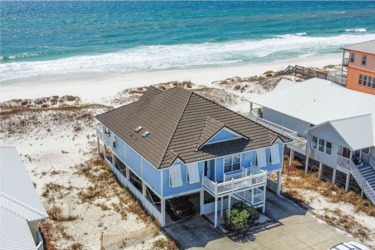 Aerial view of a two-story beach house in Gulf Shores, Alabama. The house sits atop stilts and is in the sand, less than fifty yards from the water. 