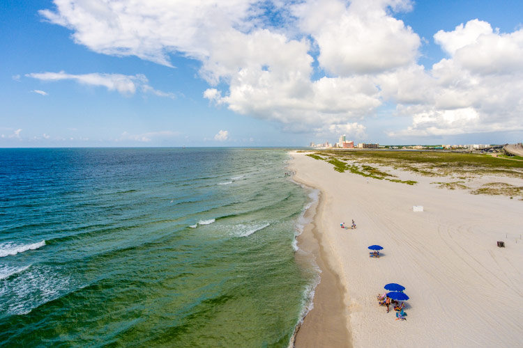 Aerial view of the coast in beautiful Orange Beach, Alabama. The gulf waters are blue and green, and a few shade umbrellas are staked into the sand where locals are enjoying a sunny summer day at the beach.