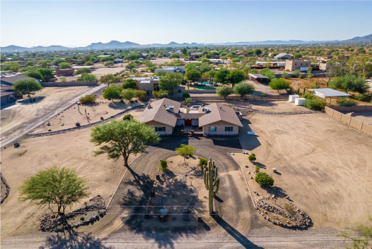 Aerial view of a residential part of Cave Creek, Arizona, on a sunny day. The large property in the foreground features a ranch-style home with mature trees and cacti.