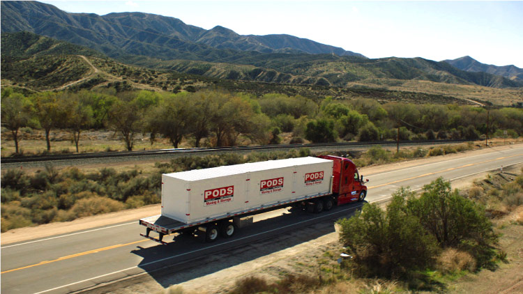 A PODS truck is transporting three PODS portable moving containers cross-country along a freeway. The truck is passing a mountain ridge and dry desert land.