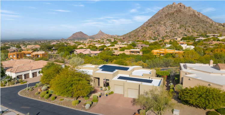 A residential neighborhood in the Troon North part of Scottsdale, Arizona. The homes are mostly made of adobe, and many properties feature gravel yards with large bushy trees. Behind the neighborhood is a tall, rocky hill. 