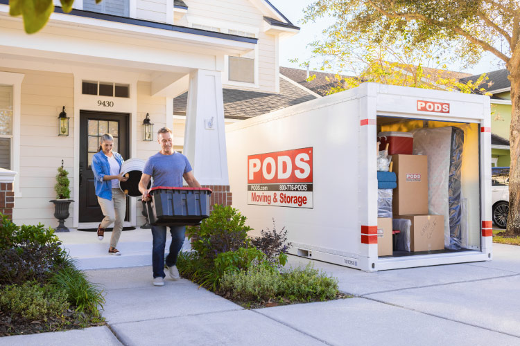 A couple is carrying things from their house to the PODS portable moving container in their driveway. The container’s door is open and it’s nearly full of boxes and furniture.