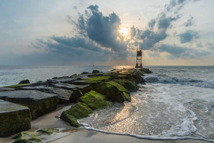 Sunrise over the ocean in Delaware. A rocky breakwater covered in green algae juts out into the ocean, hosting a tower at its end.